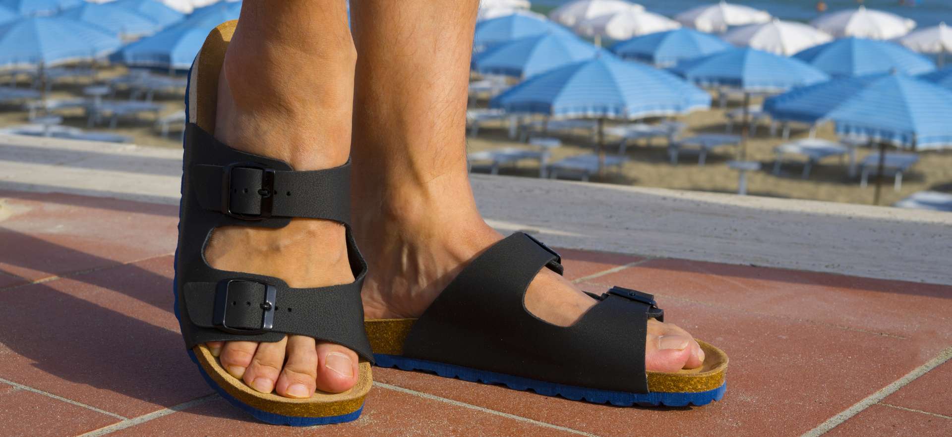 Oseba v črnih natikačah z dvema pasovoma stoji na pločniku ob plaži z ležalniki in modrimi senčniki.